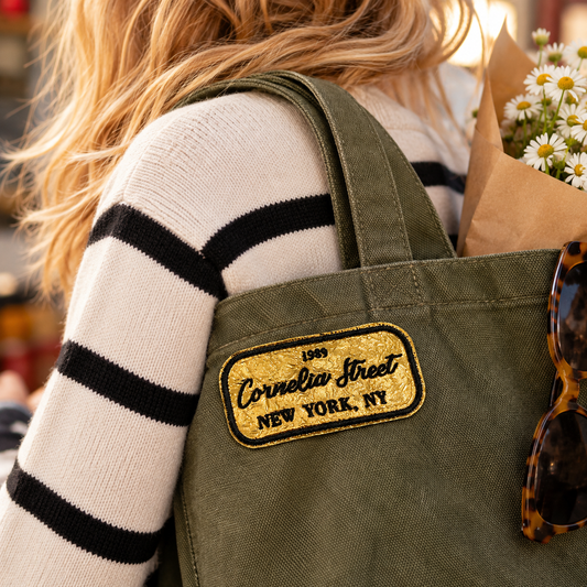 Cornelia Street gold and black embroidered patch on olive canvas tote carried over a striped sweater