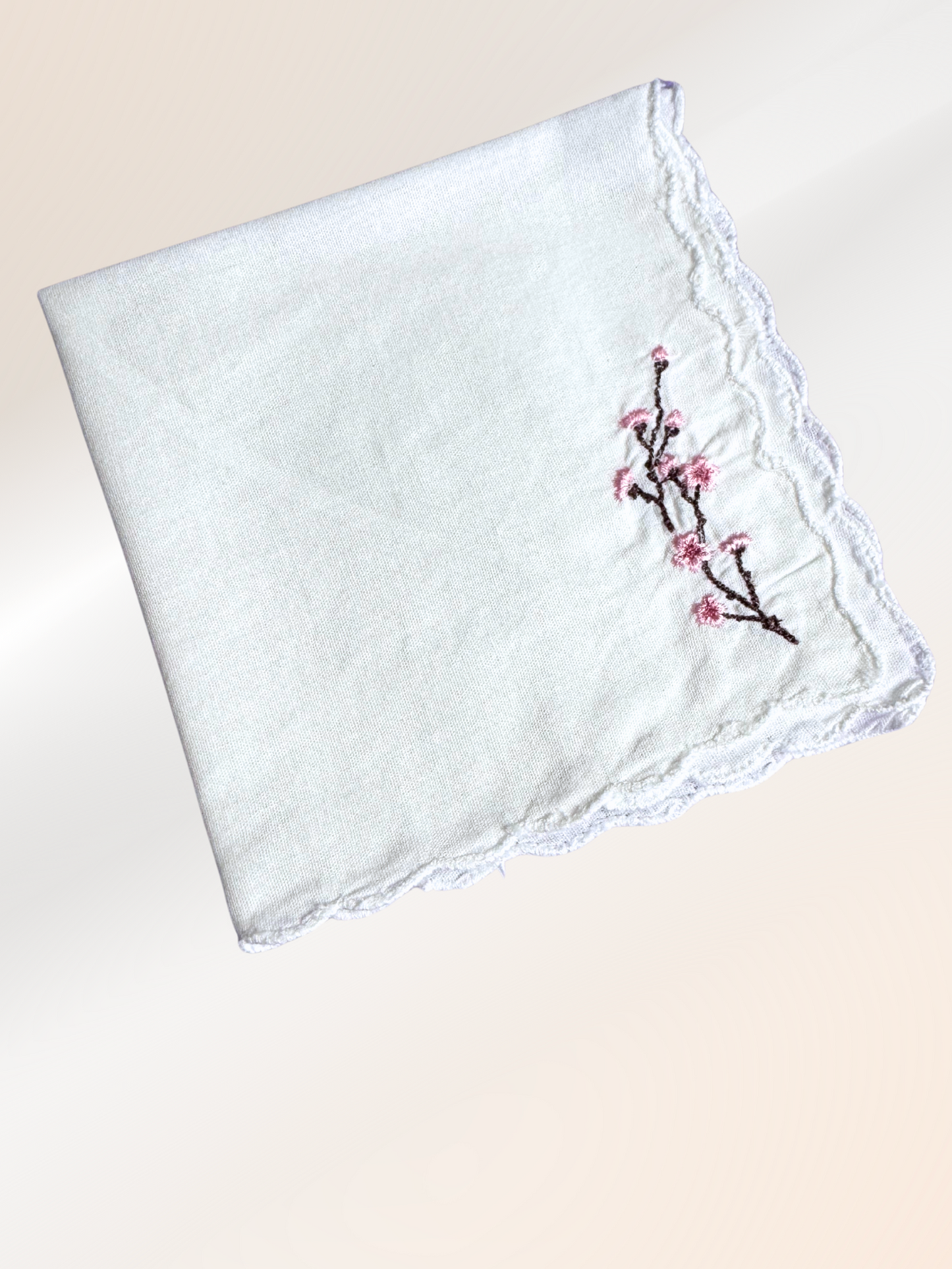 A folded embroidered handkerchief showcasing a delicate cherry blossom design stitched in pink and brown on soft white fabric, handmade in Kansas City.

