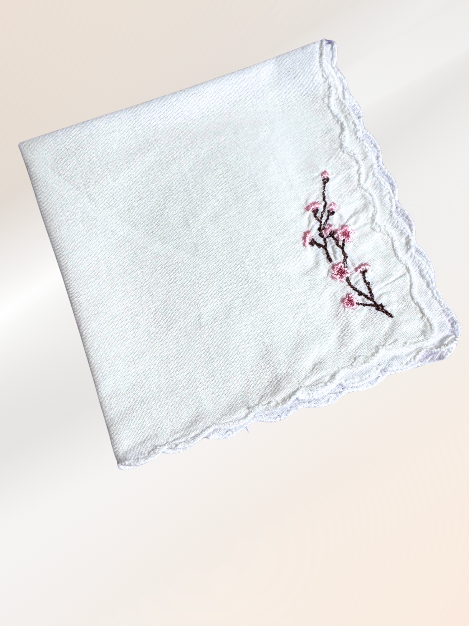 A folded embroidered handkerchief showcasing a delicate cherry blossom design stitched in pink and brown on soft white fabric, handmade in Kansas City.

