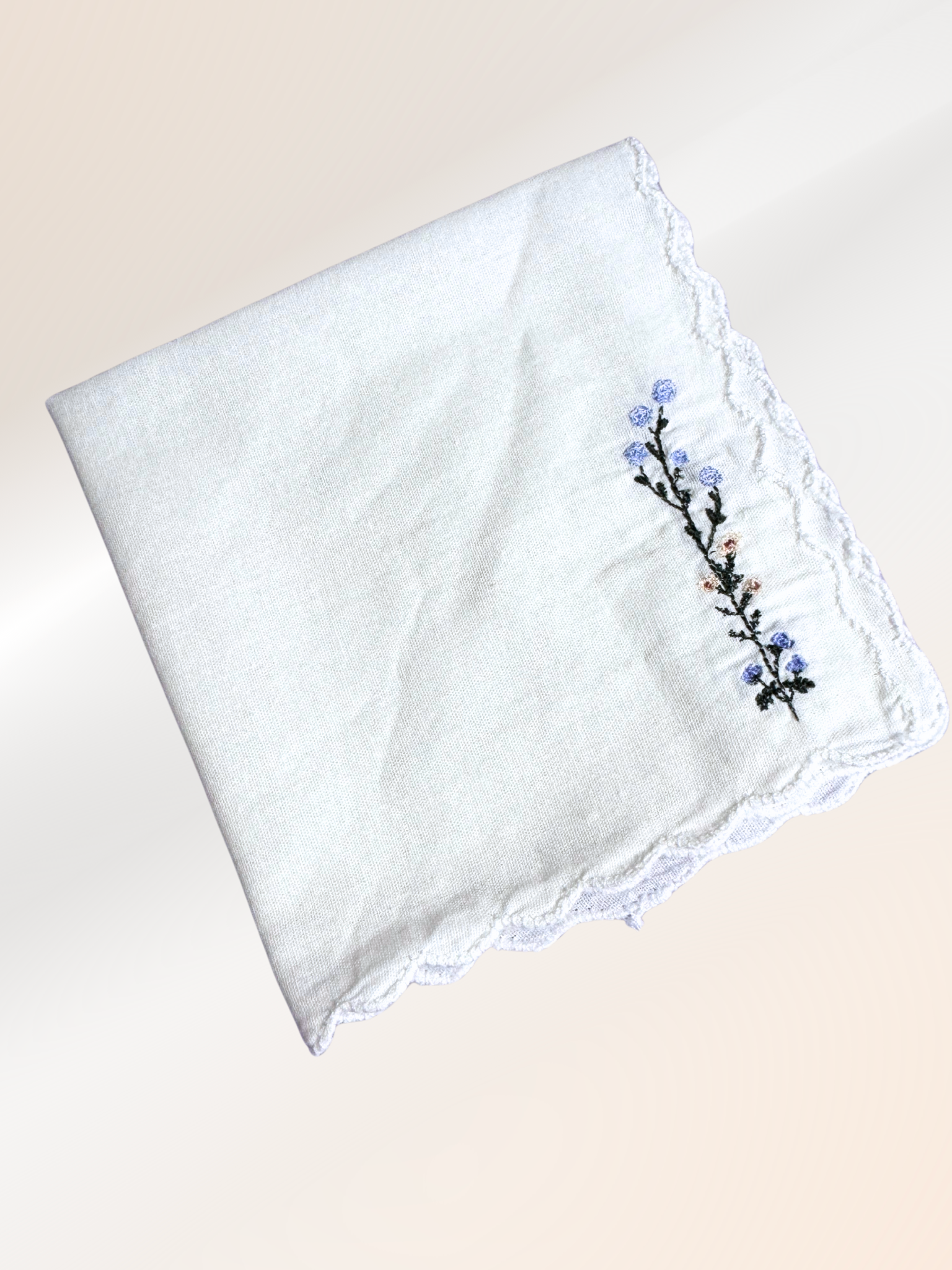 A folded embroidered handkerchief showcasing a delicate floral vine stitched in blue, peach, and green on soft white fabric, handmade in Kansas City.

