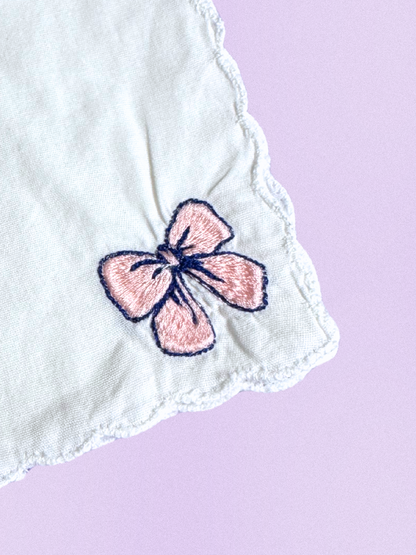 A white cotton handkerchief featuring an embroidered pink bow outlined in navy, finished with scalloped edges.
