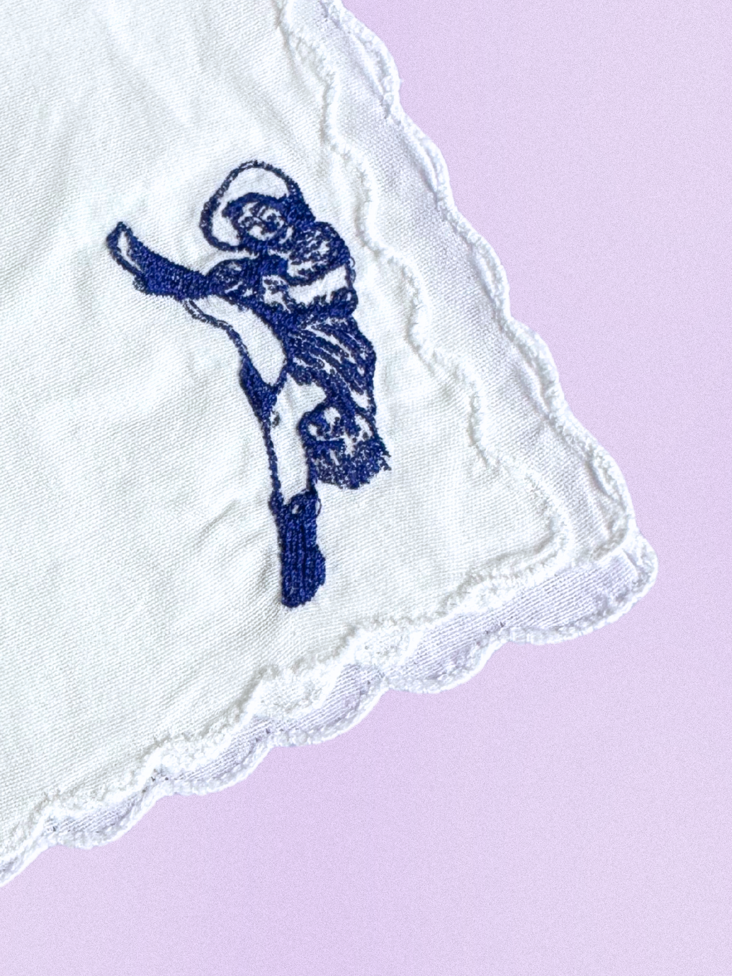 A white cotton handkerchief featuring an embroidered can-can dancer in a high-kick pose, stitched in blue thread with scalloped edges.
