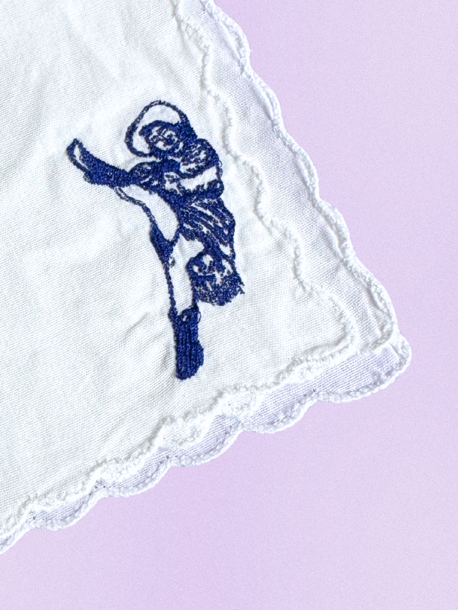 A white cotton handkerchief featuring an embroidered can-can dancer in a high-kick pose, stitched in blue thread with scalloped edges.
