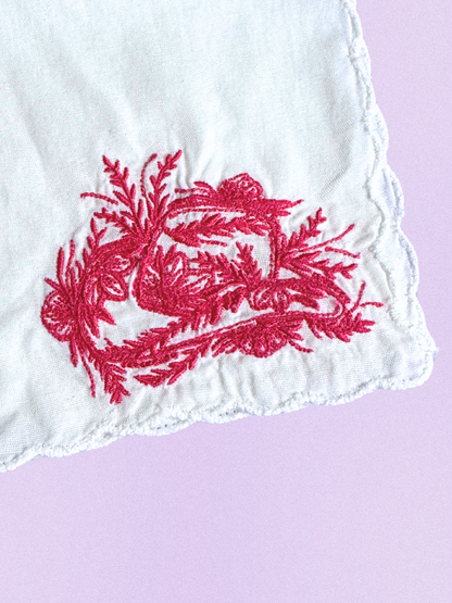 A white cotton handkerchief featuring an embroidered cowboy hat made of intricate red floral designs, finished with scalloped edges.
