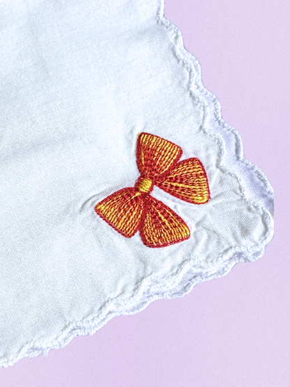A white cotton handkerchief featuring an embroidered bow in vibrant red and gold, finished with scalloped edges.

