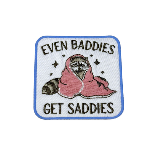 Front view of Even Baddies Get Saddies embroidered iron-on patch with raccoon wrapped in pink blanket
