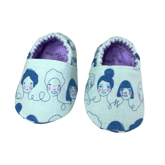 “Handmade soft sole baby shoes with blue and pink feminist icon faces print, front view, size 3-6 months”


