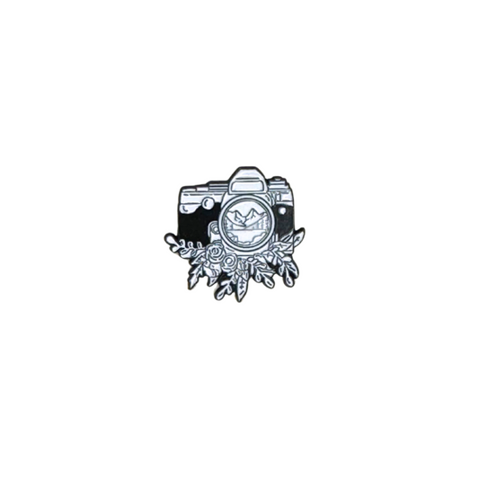 Enamel pin shaped like a camera with floral detailing and a mountain scene in the lens

