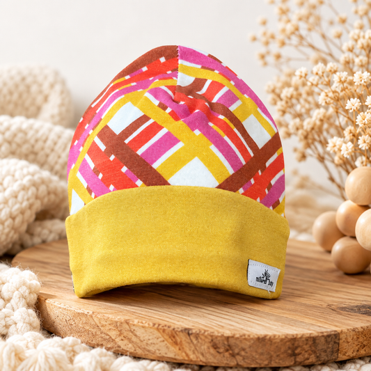 Handmade geometric pink orange brown and white baby beanie with wide mustard yellow cuff, styled on natural wood with soft cream textures