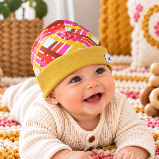 Smiling baby wearing colorful geometric knit beanie with wide mustard yellow cuff during supervised tummy time