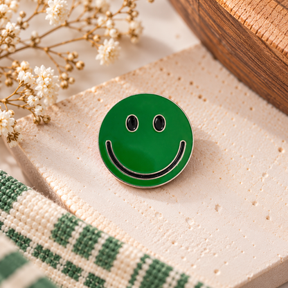 Green smiley face enamel pin on cream stone with baby's breath, wooden accent and woven textile