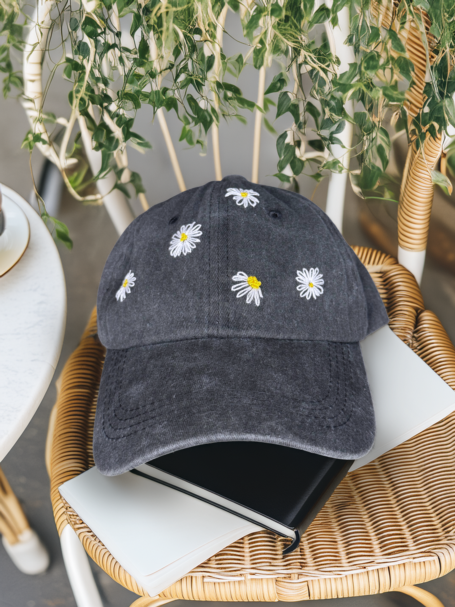 Dark gray baseball cap with white daisy flowers on a wicker chair with plants in the background
