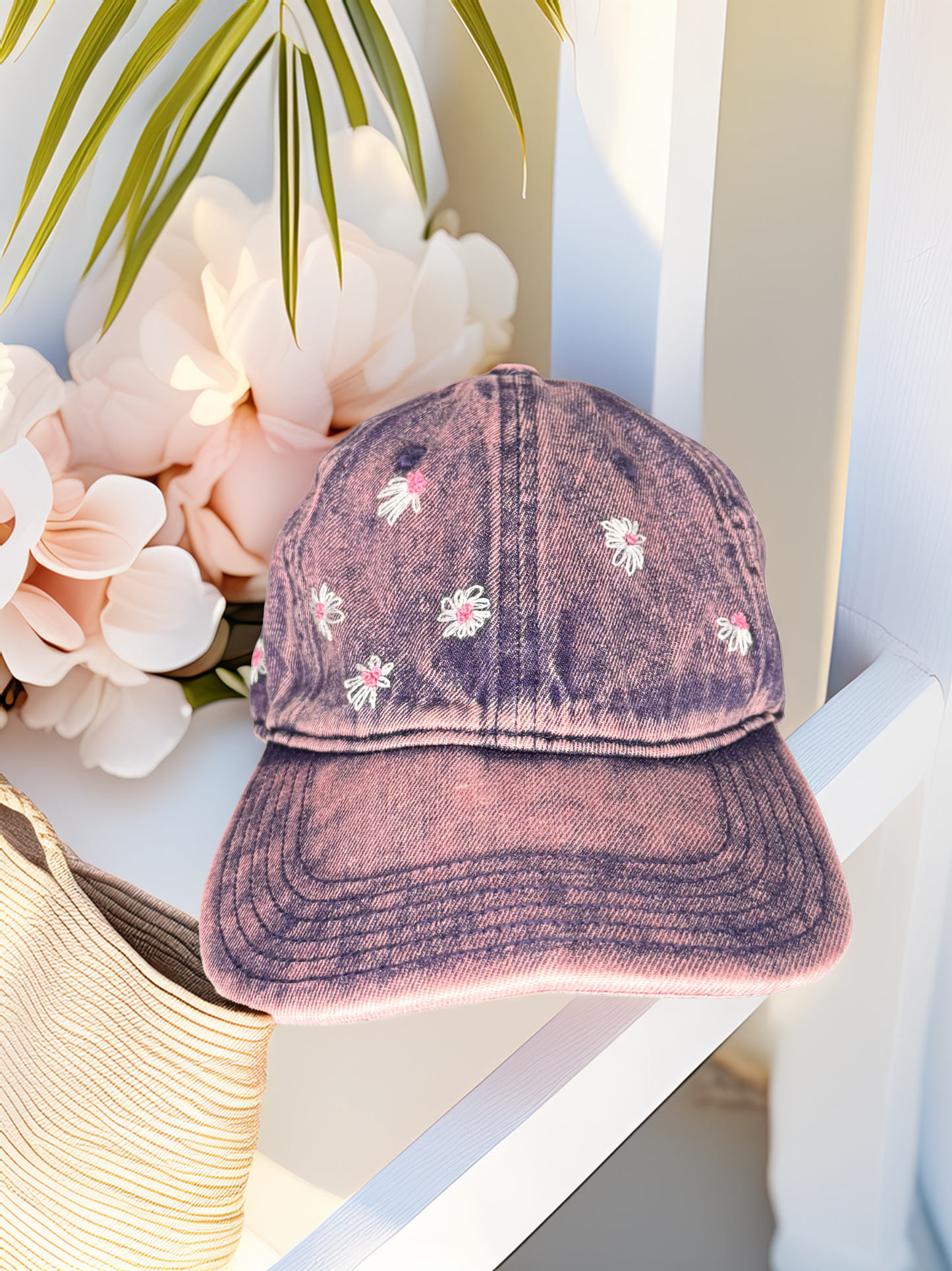 Pink distressed baseball cap with floral embroidery on a white surface with flowers and a hat.