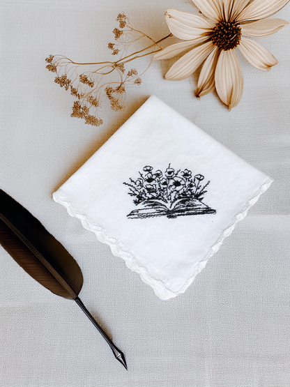 White embroidered handkerchief with book and flower design, styled with dried flowers and feather pen

