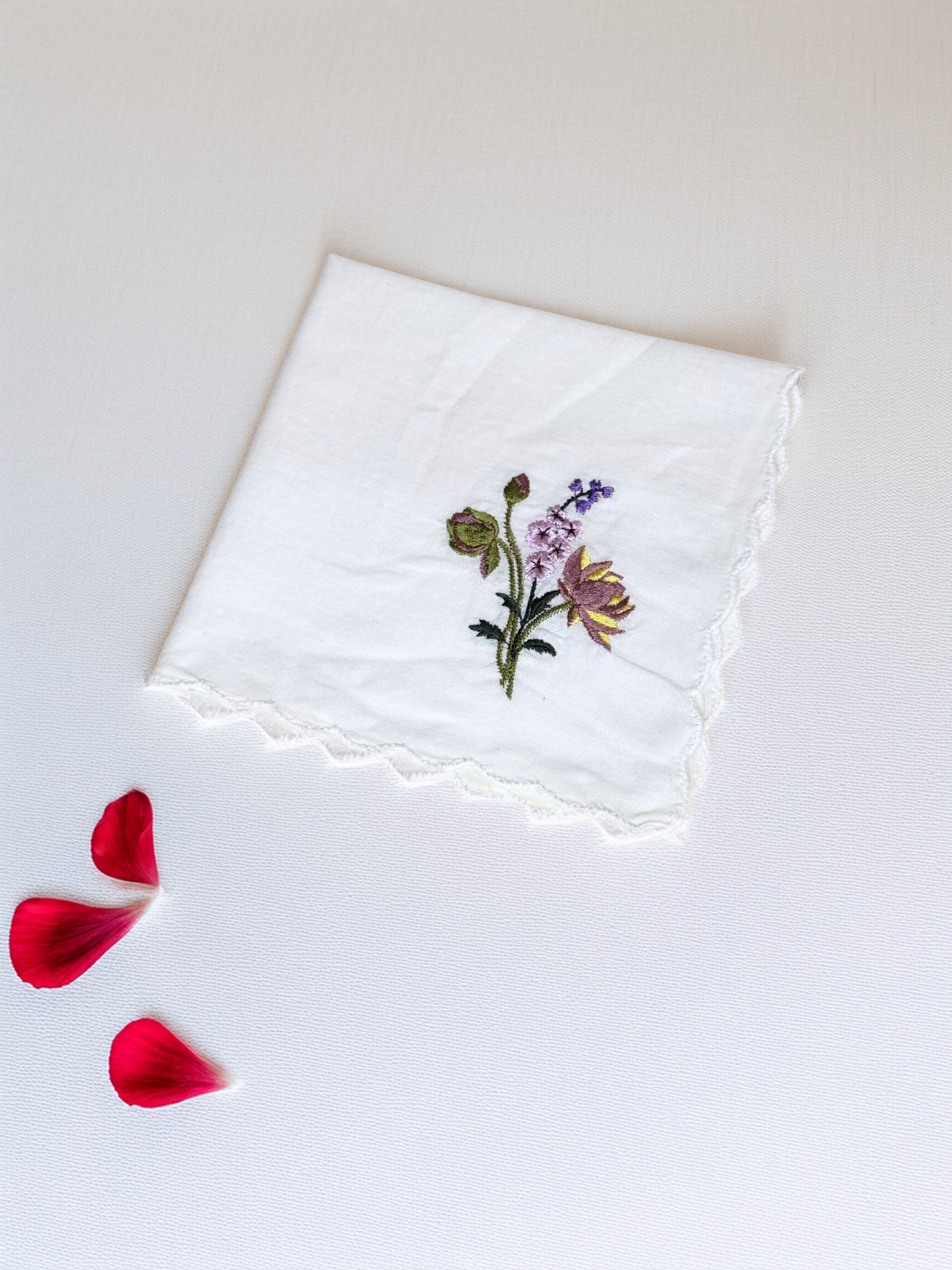 White embroidered handkerchief with purple and yellow flowers, flat lay with red rose petals

