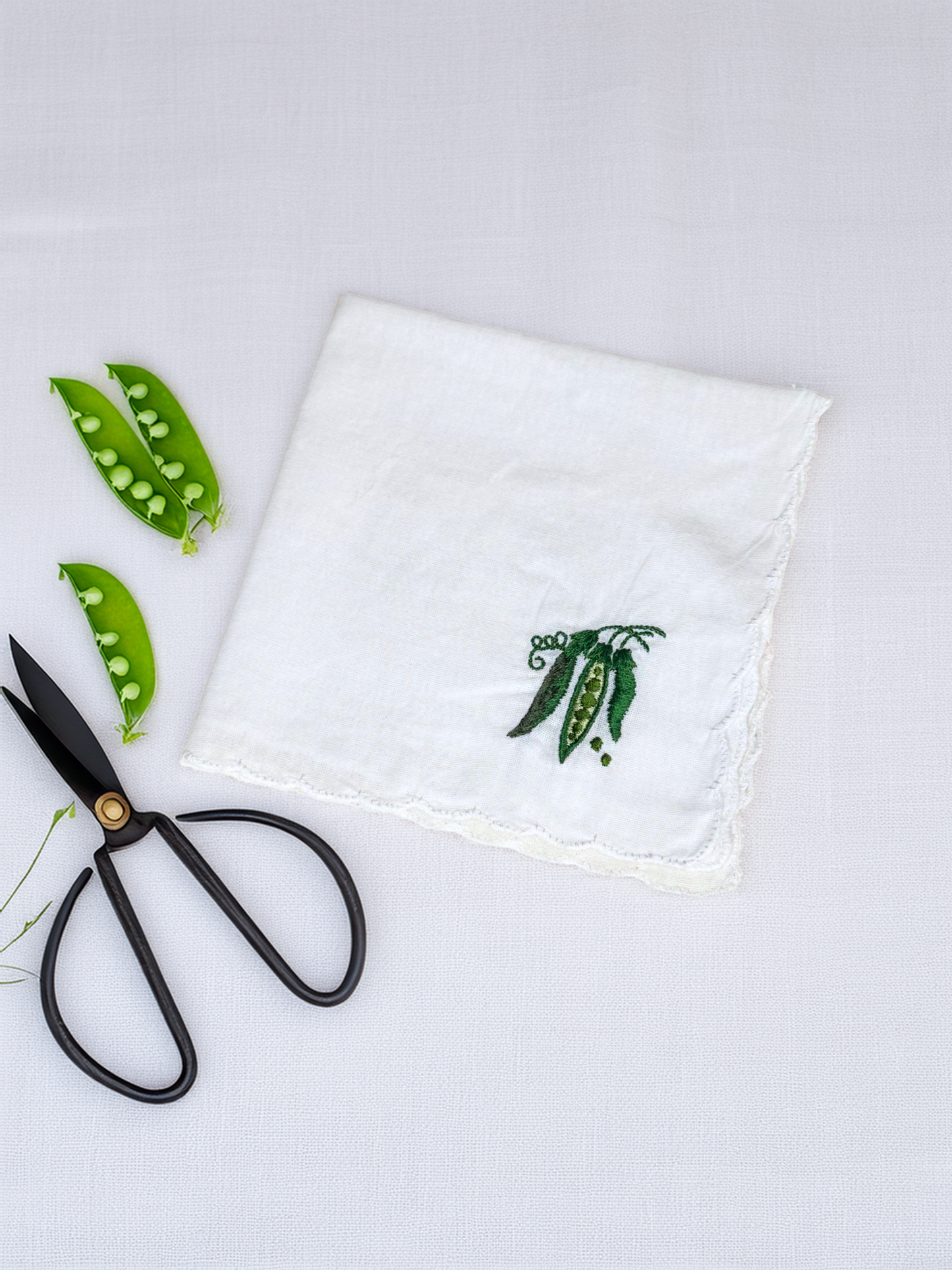 Embroidered white handkerchief with pea pod design, styled with scissors and fresh pea pods

