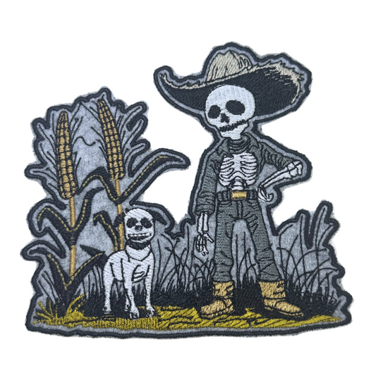 Close-up of the Harvest Guardian embroidered patch, featuring a skeleton cowboy and his dog standing in a field of cornstalks.
