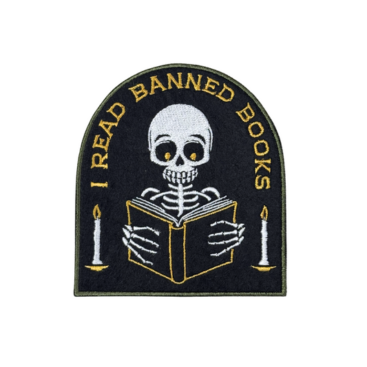 "I Read Banned Books embroidered patch featuring a skeleton reading by candlelight on a black fabric background."
