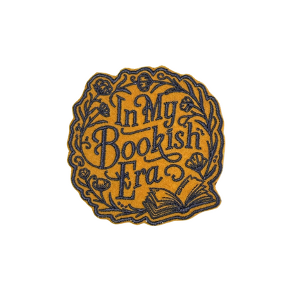 Close-up of an embroidered "In My Bookish Era" patch featuring vintage-inspired typography and floral details on a golden background.

