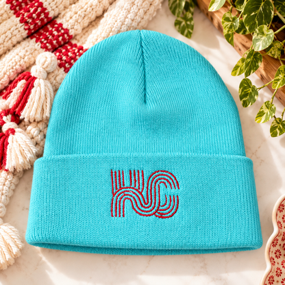 Teal knit beanie with red KC geometric embroidery on the folded cuff, styled with cream and red accents