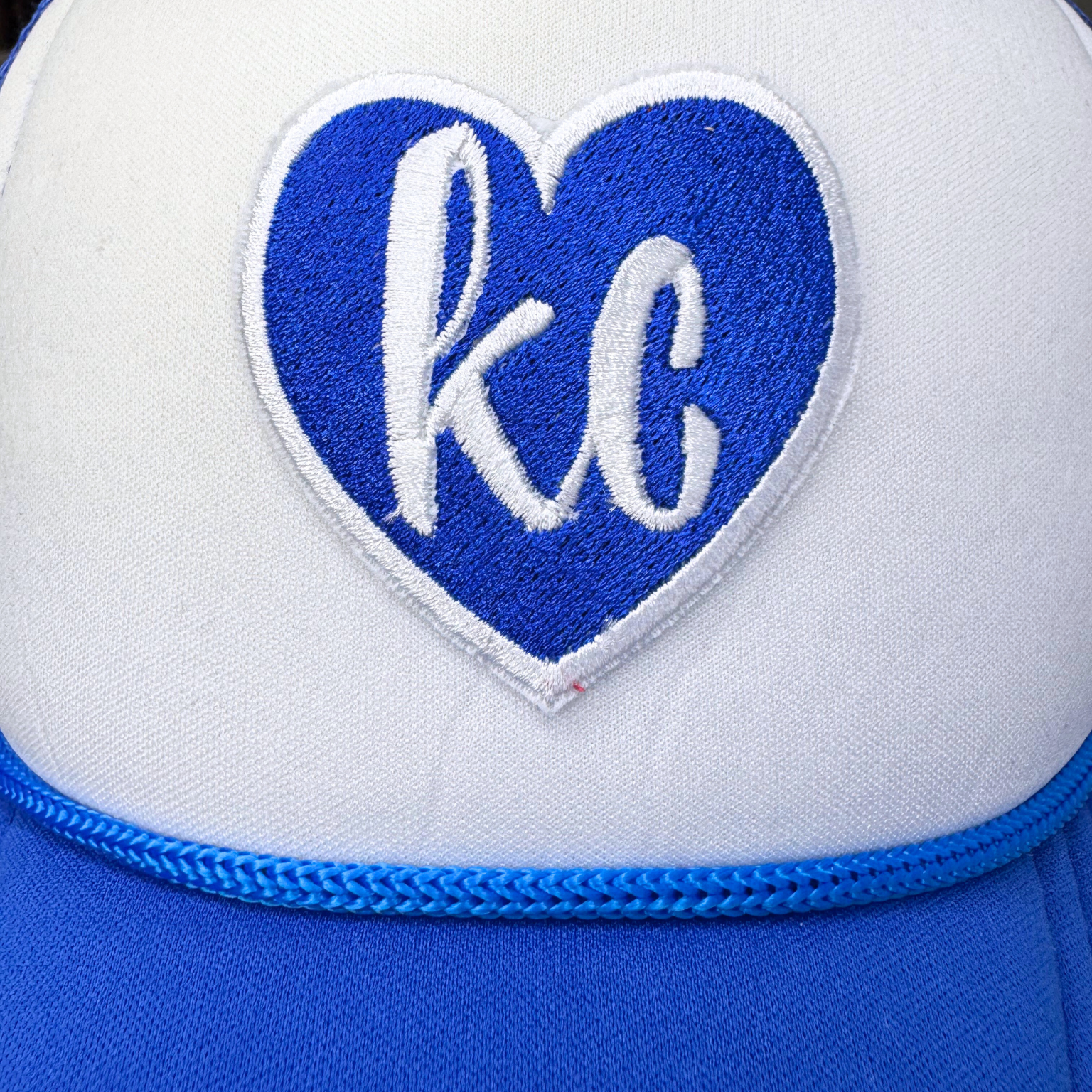 Closeup of royal blue embroidered heart patch with white lowercase "kc" on trucker hat

