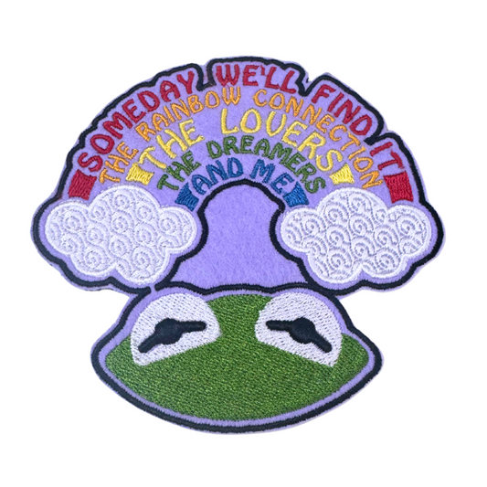 Embroidered patch featuring a green frog looking up at a rainbow sky with puffy clouds and colorful lettering.
