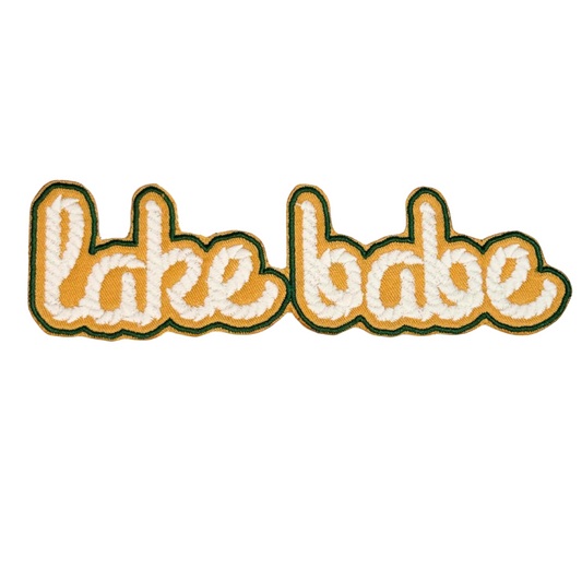 Embroidered patch that says "lake babe" in white retro lettering with gold background
