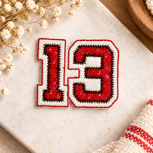 Red embroidered number 13 patch with white border and black stitching, styled on a warm cream surface