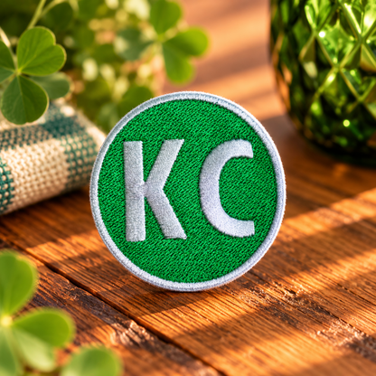 Green circular embroidered KC patch with white lettering and white stitched border, styled on warm wood with shamrocks
