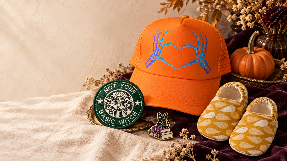 Milkweed spooky-season edit featuring an orange skeleton-heart hat, green witch patch, black cat enamel pin and mustard ghost baby shoes.