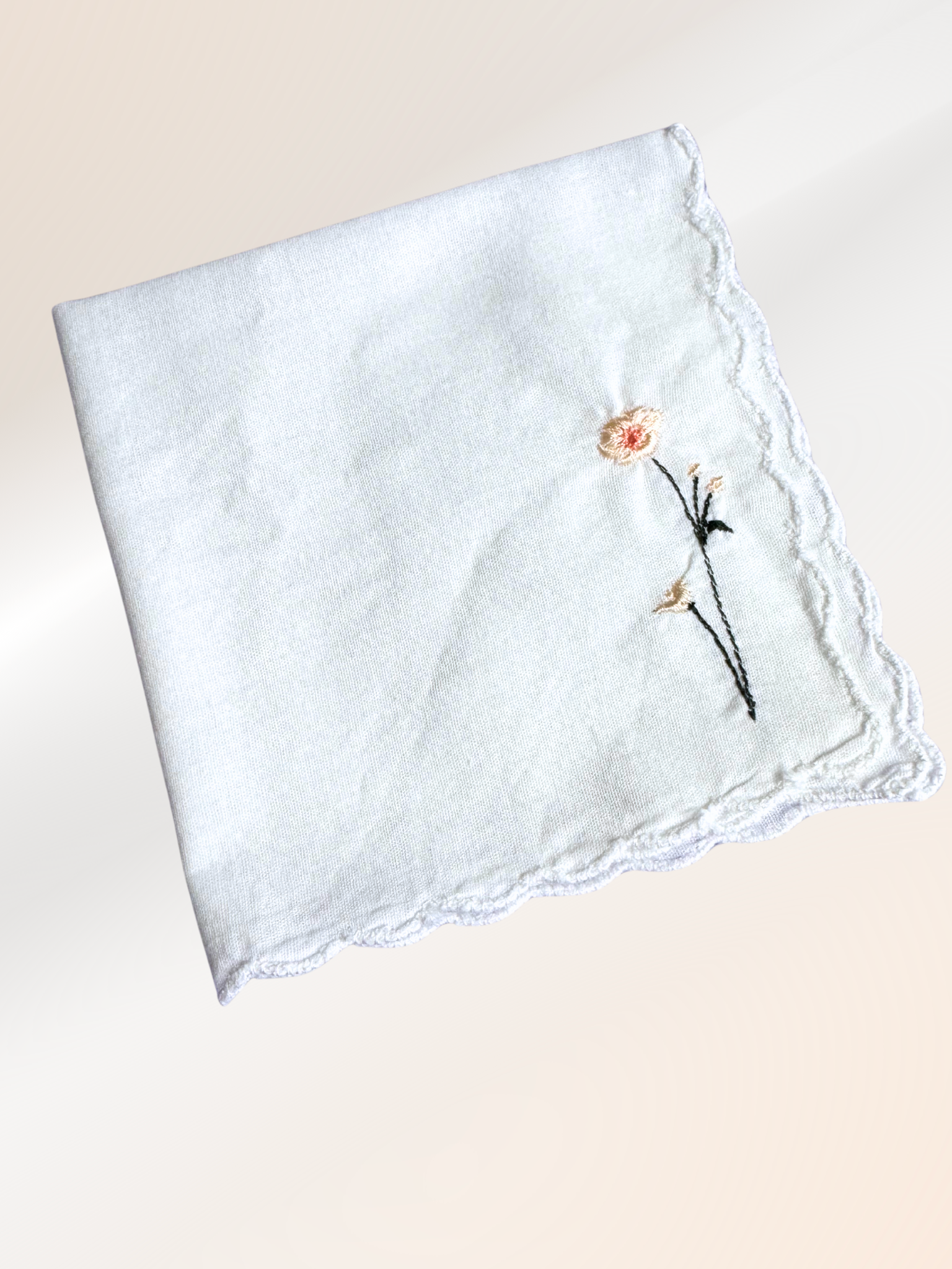 A folded embroidered handkerchief showcasing a minimalist daisy stitched in peach and green on soft white fabric, handmade in Kansas City.

