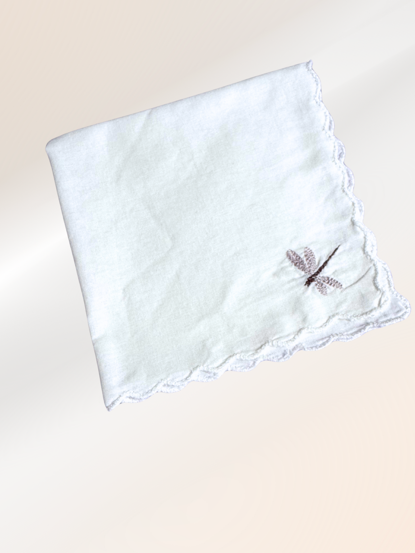 A folded embroidered handkerchief showcasing a delicate dragonfly stitched in taupe on soft white fabric, handmade in Kansas City.

