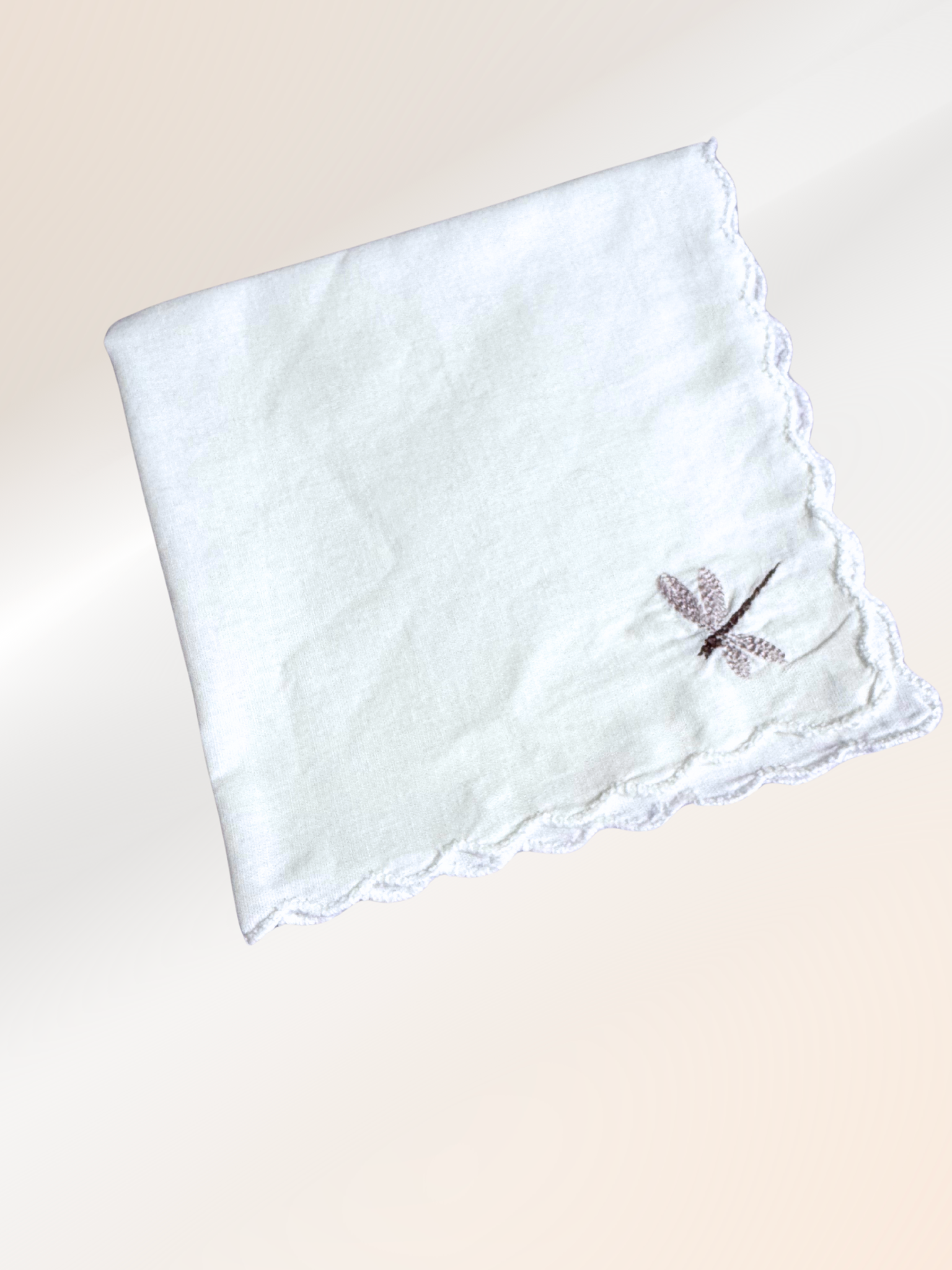 A folded embroidered handkerchief showcasing a delicate dragonfly stitched in taupe on soft white fabric, handmade in Kansas City.

