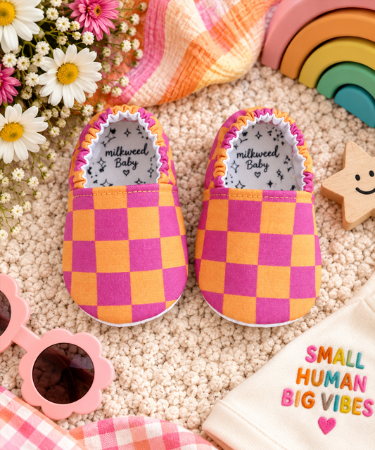 Neon Pink and Orange Checkerboard Baby Moccasins styled in a colorful Milkweed Baby lifestyle flat lay