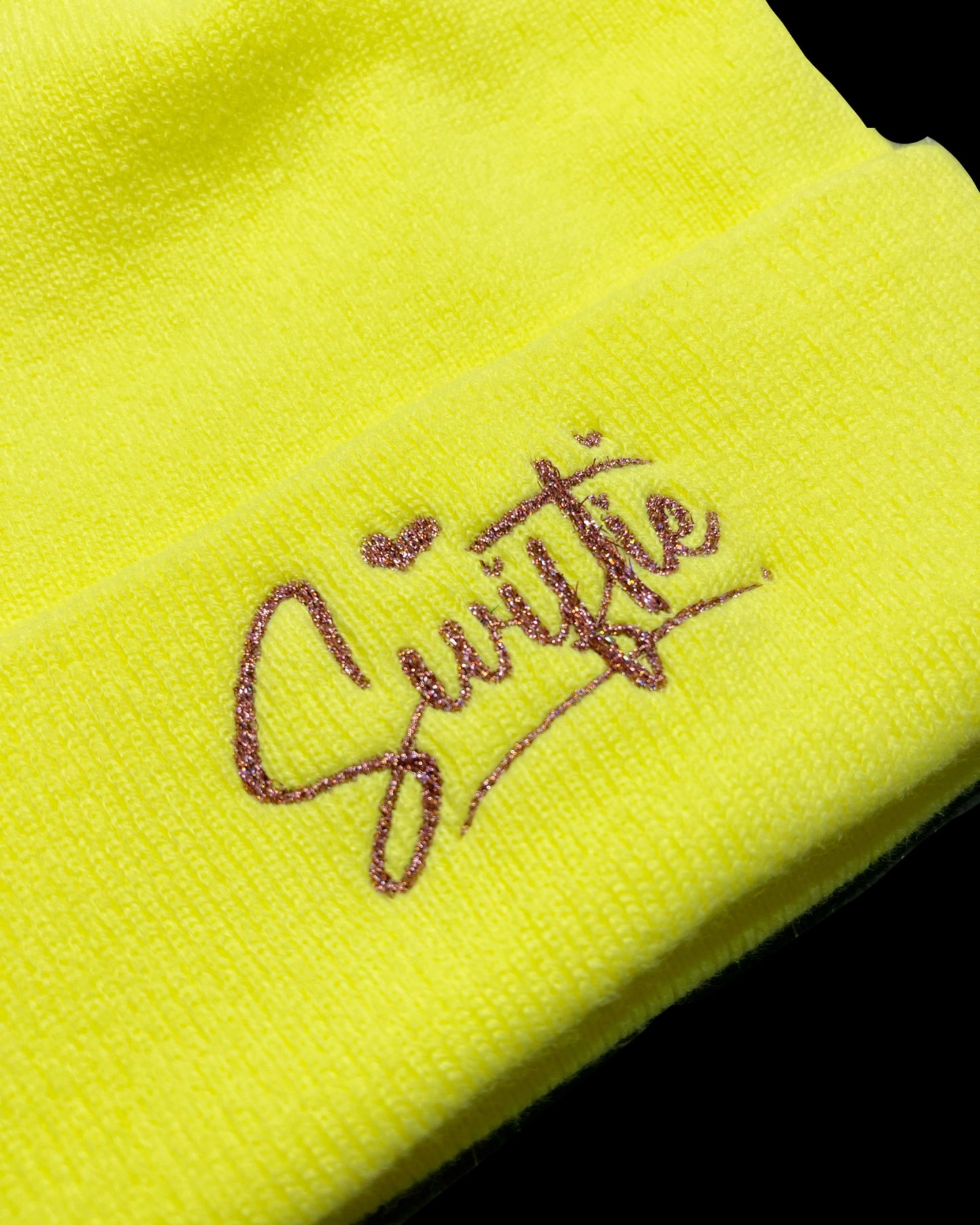 detail view of pink holographic glitter embroidery on neon yellow knit beanie word "swiftie" with hearts for the dots on the I's