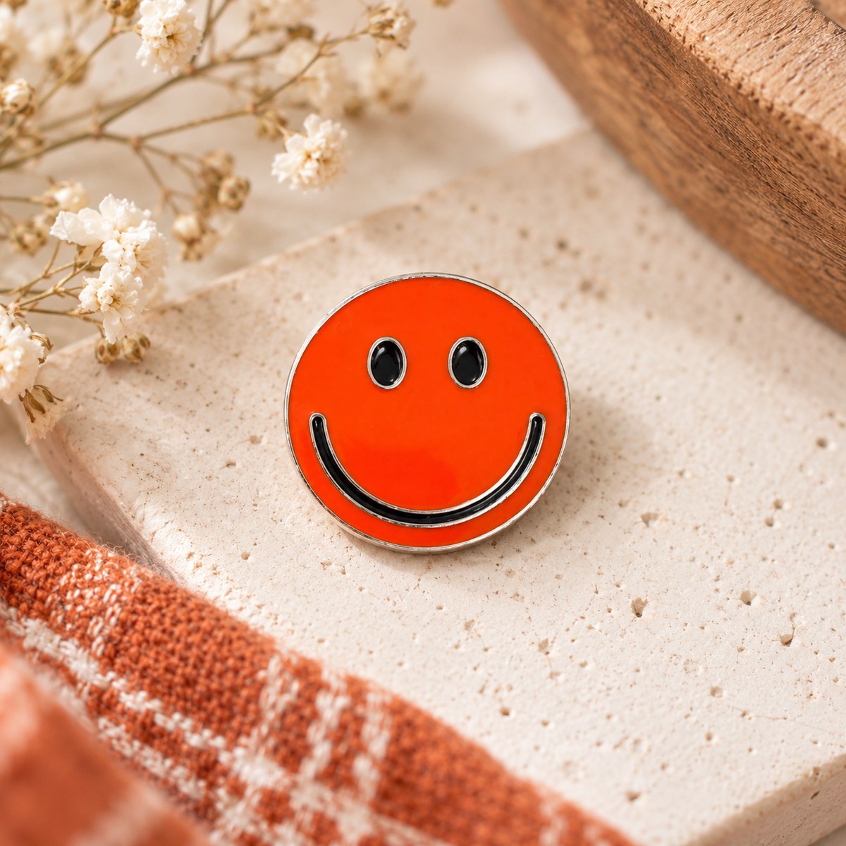 Orange smiley face enamel pin with black smile and silver outline styled on cream stone