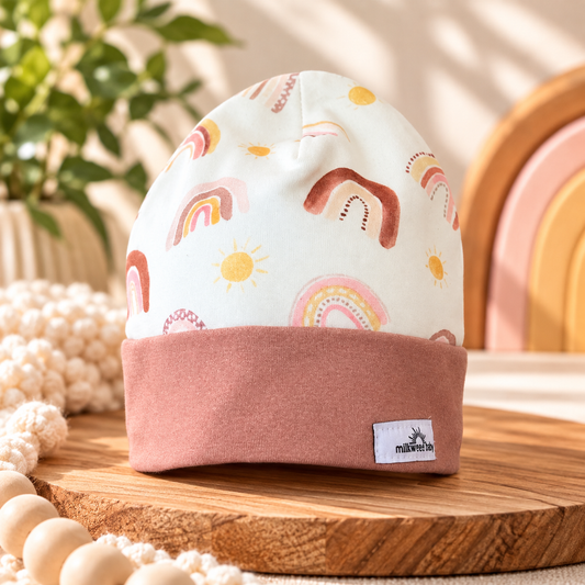 Pastel rainbow and sun organic cotton baby beanie with dusty rose cuff styled on warm wood