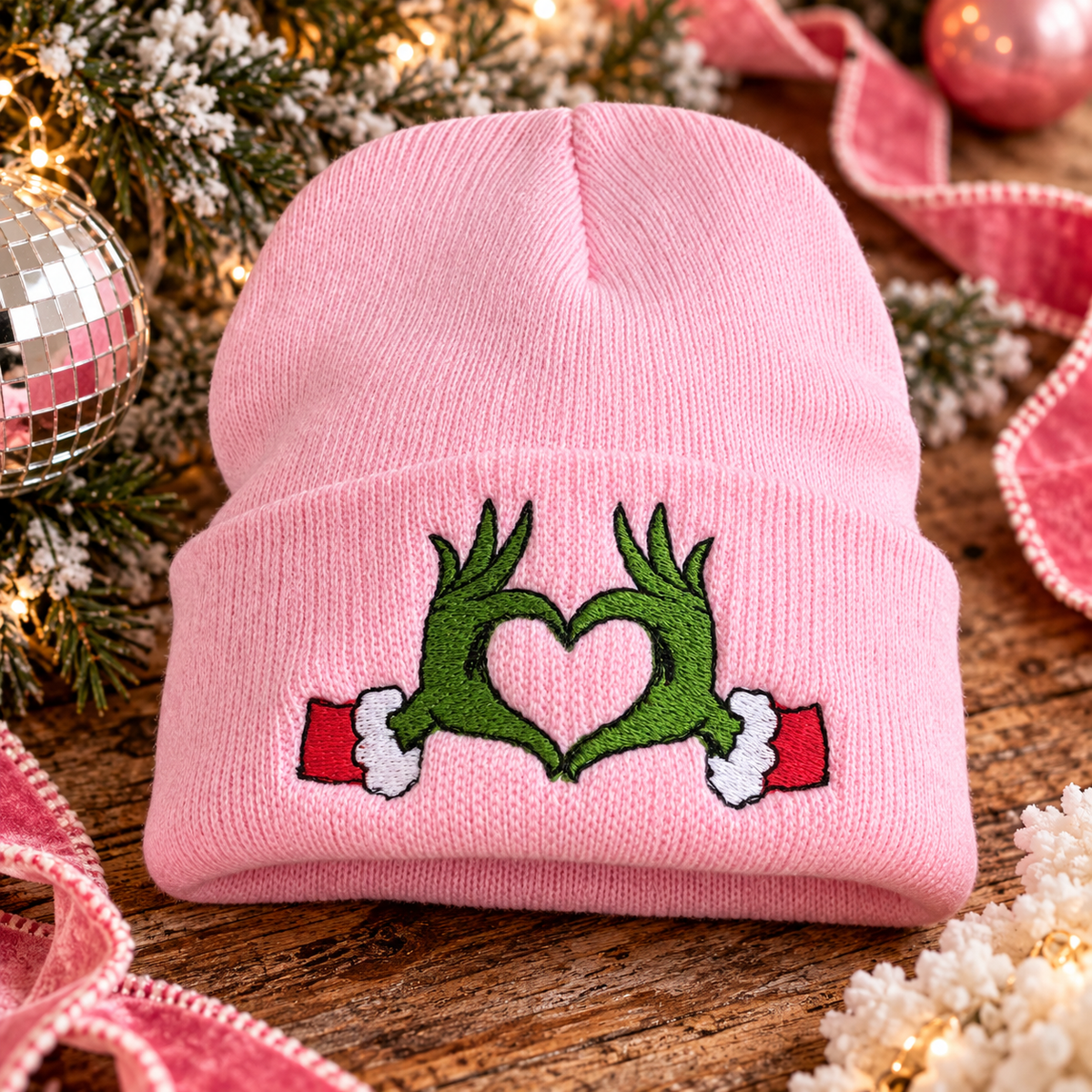 Pink embroidered Grinch heart hands beanie styled in a festive holiday scene.