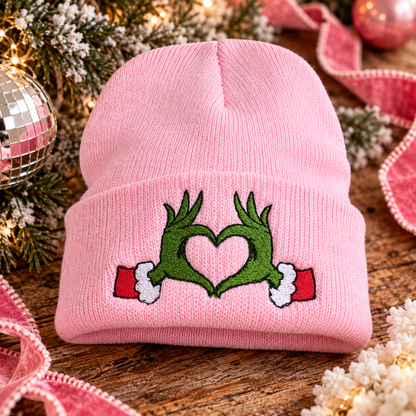 Pink embroidered Grinch heart hands beanie styled in a festive holiday scene.