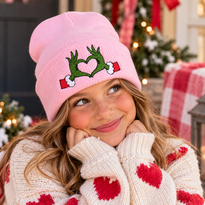 Youth wearing pink embroidered Grinch heart hands beanie in a festive holiday setting.