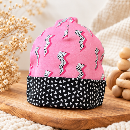 Pink organic cotton baby beanie with spotted lightning bolts and black-and-white triangle cuff, styled with natural wood and cream textures