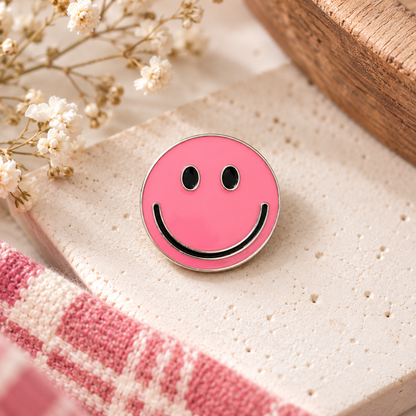 Pink smiley face enamel pin styled on cream stone with flowers