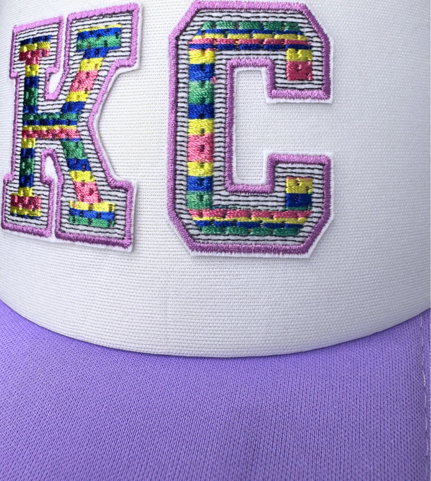 Closeup of embroidered rainbow-striped "KC" patch on lilac trucker hat

