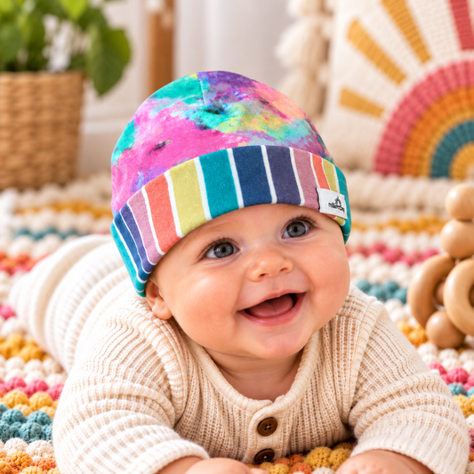Smiling baby wearing rainbow tie-dye beanie with full-width multicolor striped cuff, styled in a cozy nursery