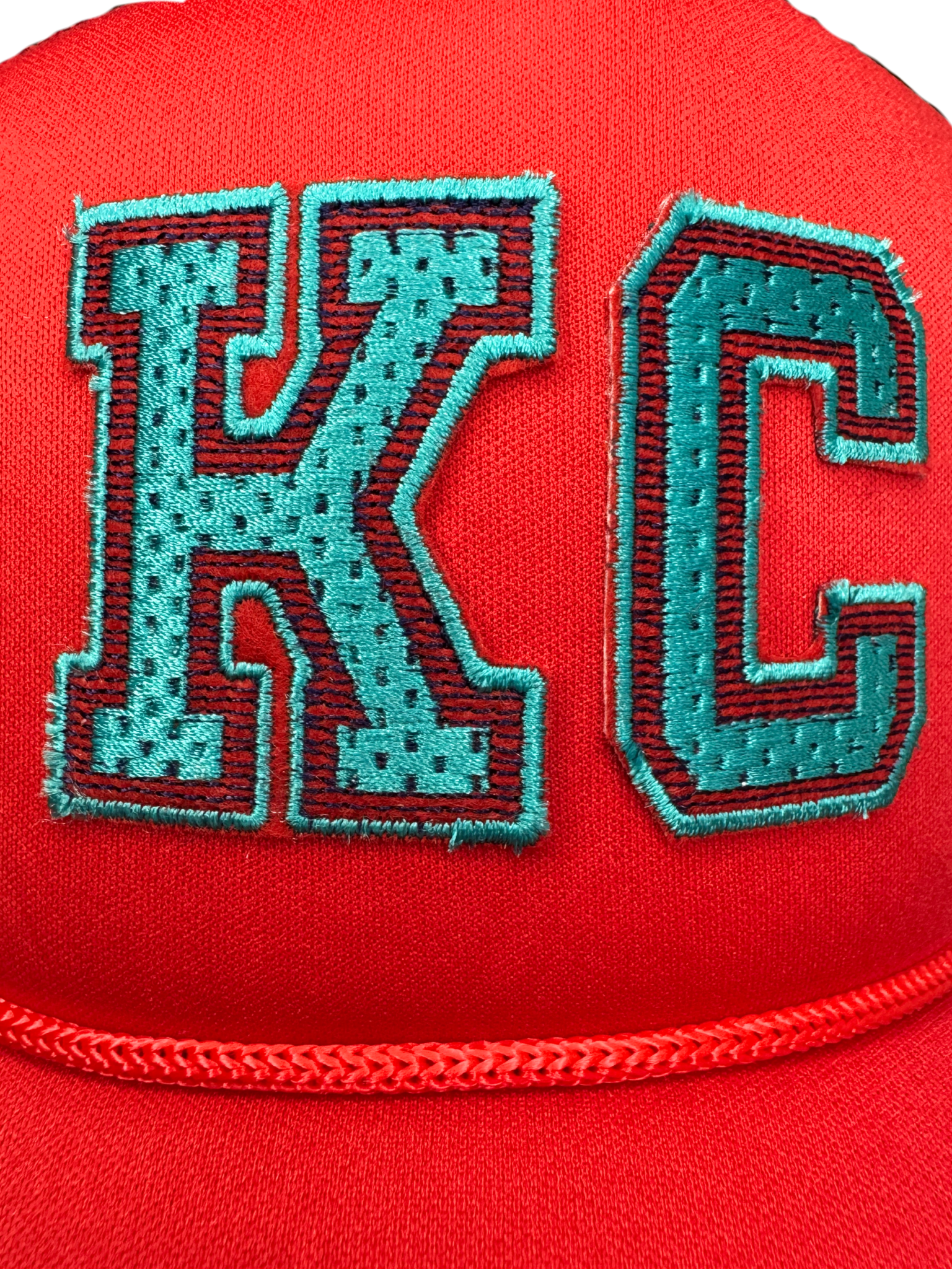 Closeup of varsity-style KC patch in teal embroidery on red trucker hat

