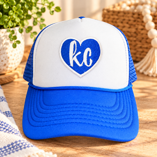 Royal blue and white KC heart trucker hat with embroidered heart patch, styled on a warm wood tabletop