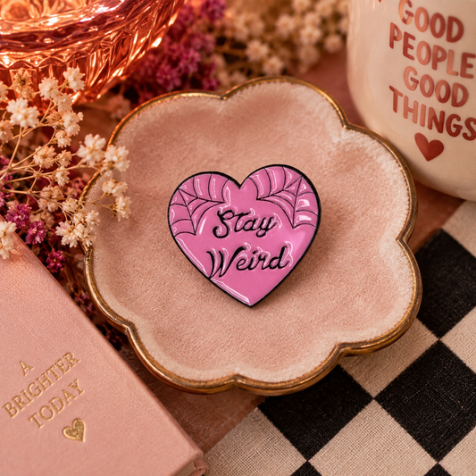 Stay Weird pink heart enamel pin styled in a warm boutique flat lay
