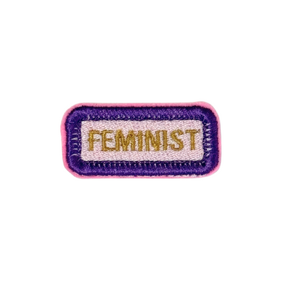 "Embroidered rectangular patch featuring the word 'FEMINIST' in gold on a white background, outlined in purple and pink, representing suffragette colors."
