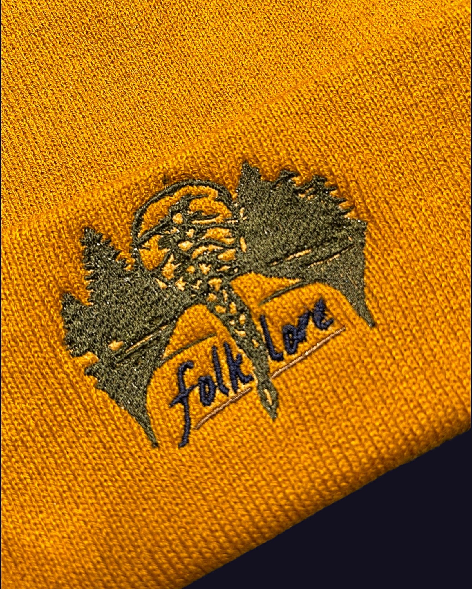 closeup of embroidery in olive green of forest and back of Taylor swifts head with hair in a braid and the word folklore underlined on a golden yellow knit beanie