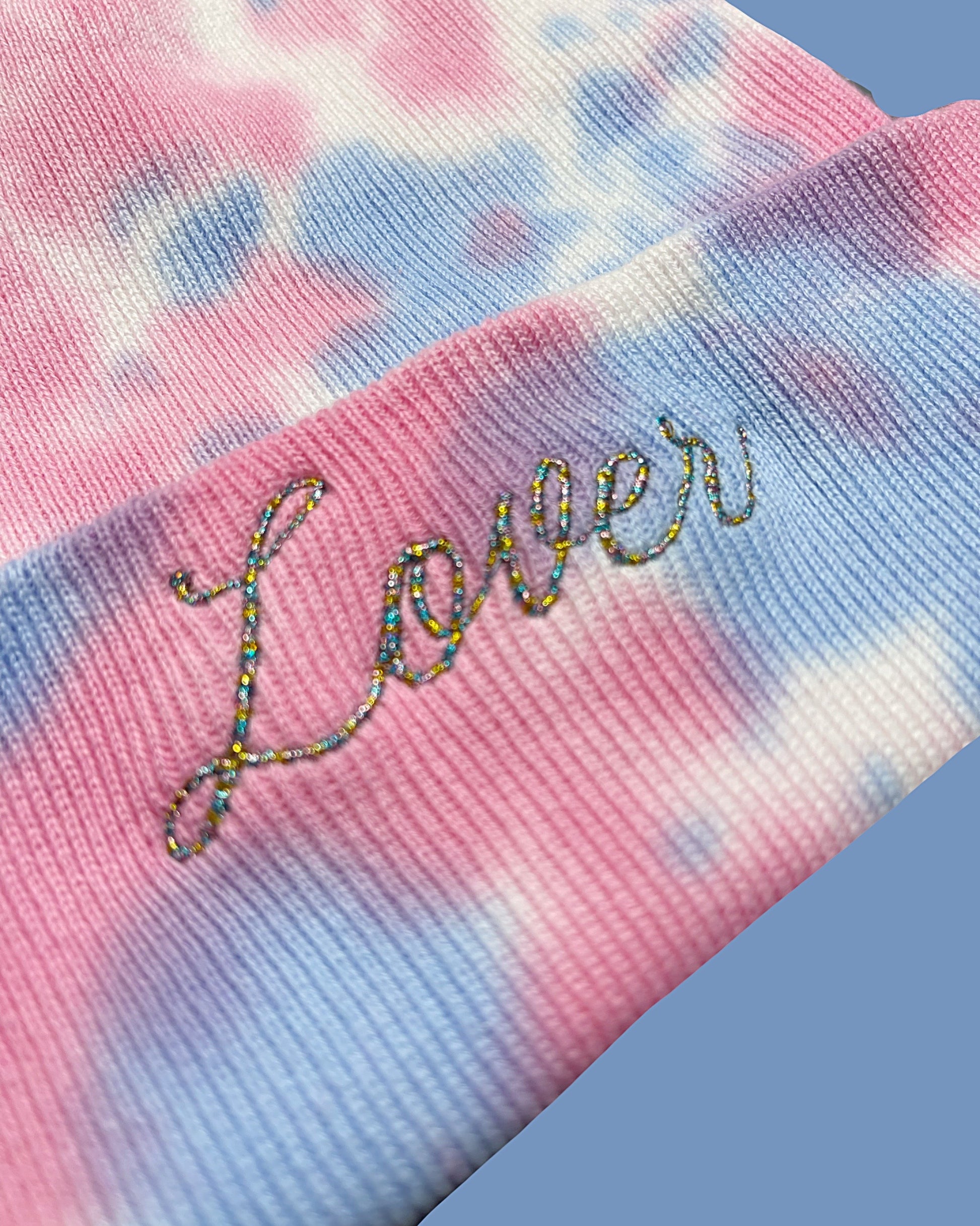 detail view of pink and blue tie dye knit beanie embroidered in variegated metallic thread with "LOVER" like the cover to the Taylor Swift Album of the same name